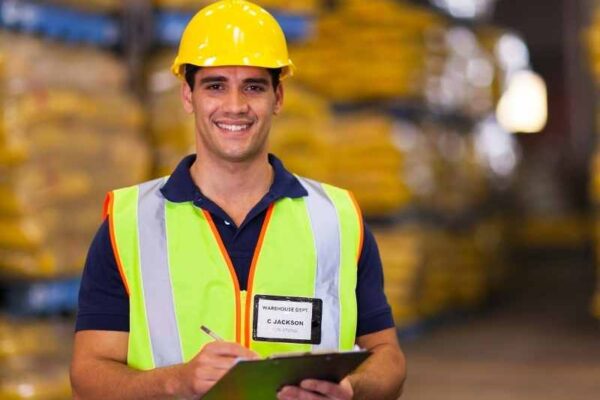 Trabajador de almacén con chaleco y casco revisando inventario en un centro de distribución.