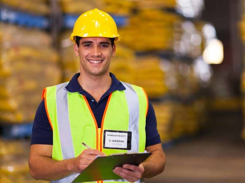 Trabajador de almacén con chaleco y casco revisando inventario en un centro de distribución.