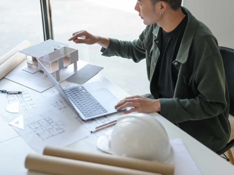 Fotografía de un plano arquitectónico extendido sobre una mesa de metal, con un casco de construcción y una tablet al lado. Luz de estudio, enfoque profesional.